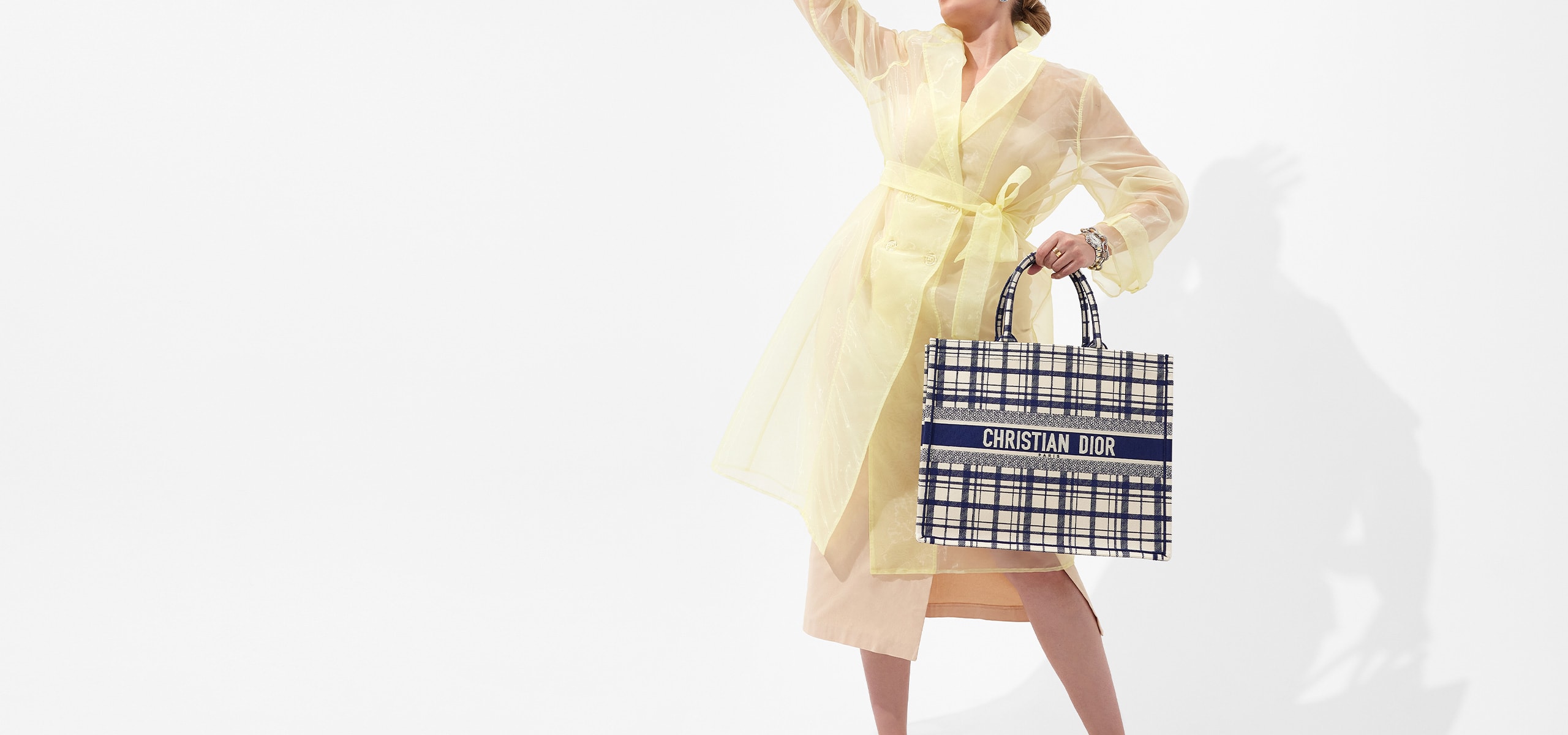 woman in sheer yellow trench coat holding a navy Christian Dior book tote